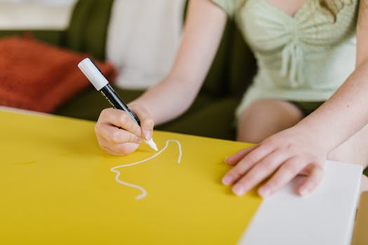 A child in a green dress drawing with a marker on yellow paper indoors.