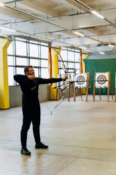 A man practicing archery indoors, aiming at a bullseye target with focused precision.