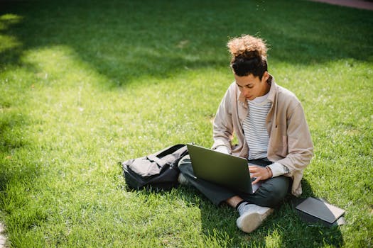 A young man sits on the grass studying with a laptop in a park setting.