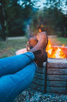 Unrecognizable tourist in jeans and boots chilling with legs crossed by bright bonfire in big fire bowl in forest on blurred background