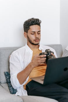 Young bearded Hispanic male learning to use professional photo camera while watching video tutorial on laptop during free time at home